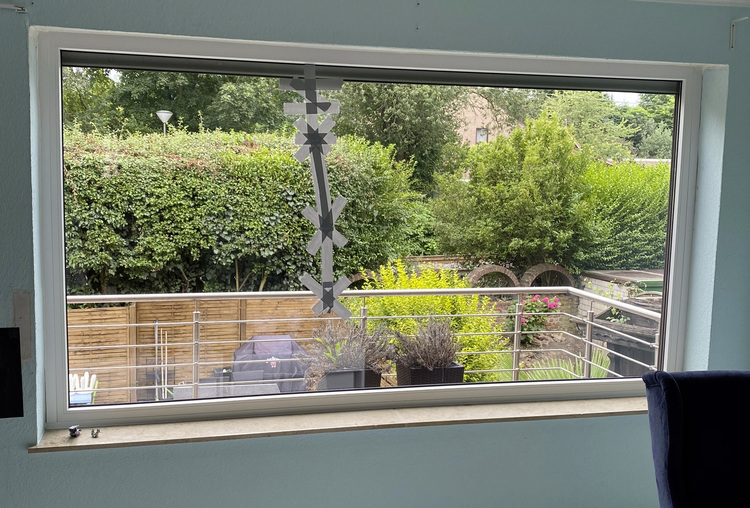 Großes Fenster mit Blick auf eine begrünte Terrasse, einen Grill und eine Hecke; am Glas hängen Schneeflocken-Dekorationen. Der Fokus liegt auf der Gebäudehülle und dem Außenbereich.