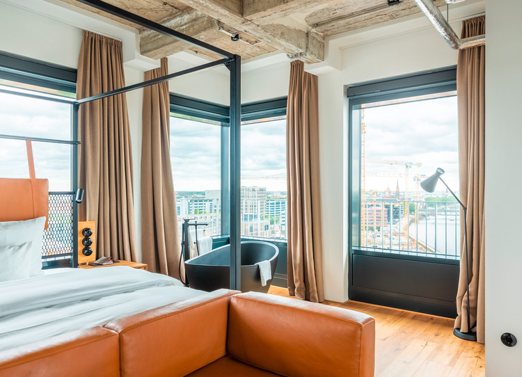 Ein modernes Hotelzimmer mit bodentiefen Fenstern, einer schwarzen Badewanne und einer Sichtbetondecke als Beispiel für energieeffiziente Gebäudetechnik.