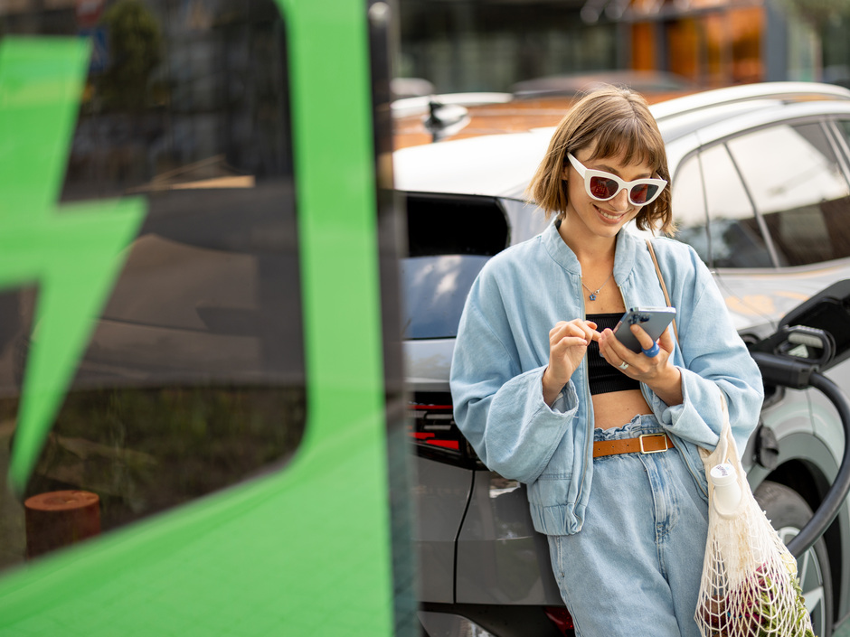 Junge Frau mit einem Einkaufstasche steht vor Ladesäule