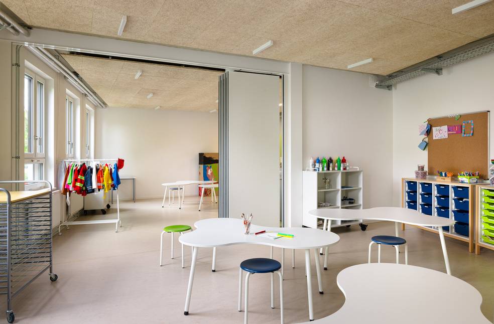 Alt-Text: „Heller Seminarraum mit wellenförmigen Tischen, Hockern, farbigen Lehrmaterialien und Kunstwerken an den Wänden als Beispiel für moderne Innenraumgestaltung in Bildungsgebäuden.