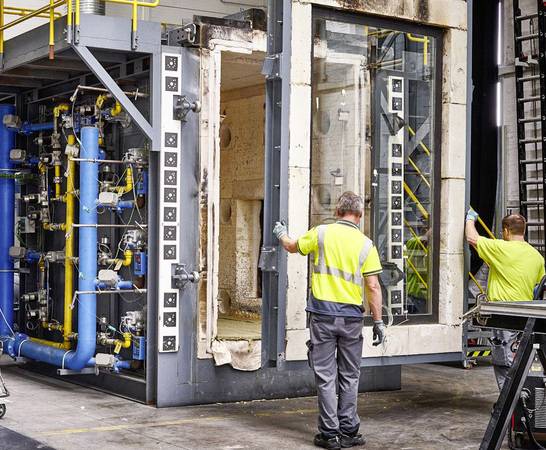 Das vorbereitete Bauteil mit den Abmessungen von 1,50 × 2,50 m – Rahmen und Brandschutzglas – wird mit dem Hallenkran vor dem Ofen in Position gebracht. Dann wird der Brandraum mit dem Bauteil verschlossen.  Das vorbereitete Bauteil mit den Abmessungen von 1,50 × 2,50 m – Rahmen und Brandschutzglas – wird mit dem Hallenkran vor dem Ofen in Position gebracht. Dann wird der Brandraum mit dem Bauteil verschlossen.