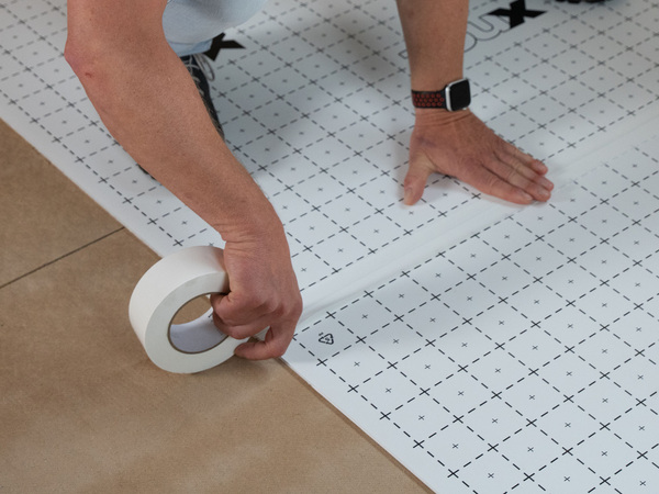 Eine Fachkraft befestigt mit Malerkrepp eine weiße, rasterförmige Dämm- oder Messmatte auf dem Boden zur Vorbereitung gebäudetechnischer Arbeiten.