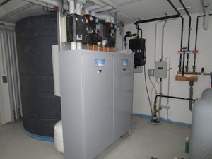 Hier ist im Hintergrund ein 4200-Liter-Pufferspeicher zu sehen. Er ist verbunden mit einer Luftwärmepumpe, die außerhalb des Hauses arbeitet. Der „Wärmemanager“ im Vordergrund sorgt für ein optimiertes Zusammenspiel der Komponenten, welches sich nach dem jeweiligen Angebot an solarer Energie und dem aktuellen Bedarf an Wärme und Haushaltsstrom richtet. Flexible Stromtarife können ebenfalls berücksichtigt werden, sofern sie vom Versorger bereits angeboten werden.